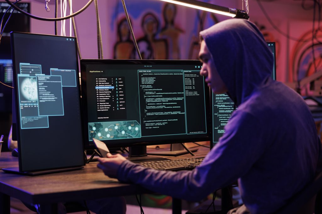 Asian hacker working on multiple monitors displaying code and applications, illustrating malware analysis and virus removal services by Bizup IT in Murray Bridge.