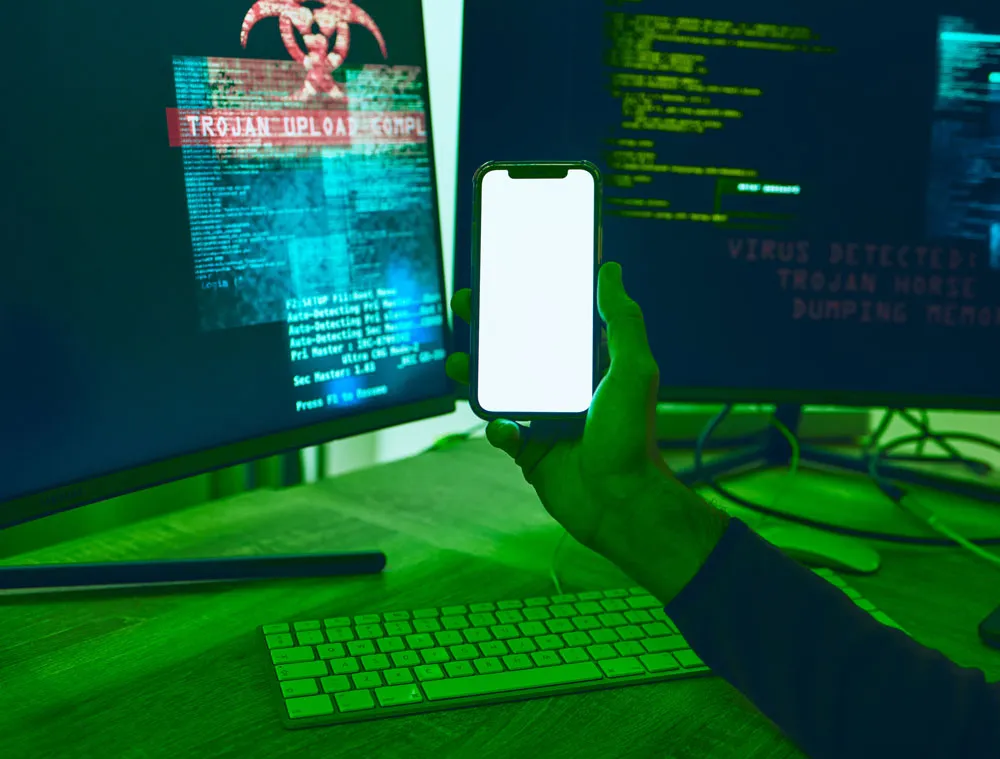 Hand holding a smartphone in front of dual computer monitors displaying virus alerts and malware warnings, illustrating phone and computer virus repair services in Murray Bridge by Bizup.