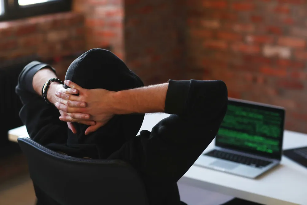 Person in a black hoodie sitting at a desk with hands clasped behind their head, looking at a laptop displaying green code, symbolizing computer security and hacking concerns relevant to BizUp's repair services in Murray Bridge.