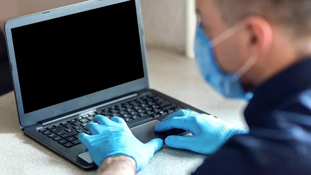 Person wearing blue gloves and a face mask working on a laptop, representing professional computer repair services in Broken Hill.