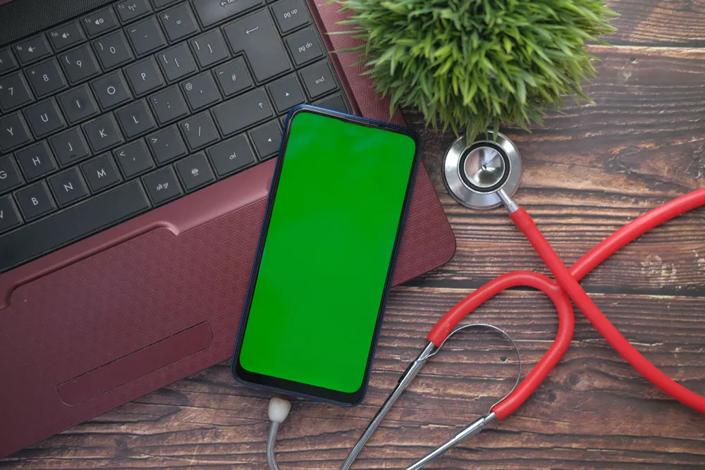 Smartphone with green screen on laptop keyboard, stethoscope and potted plant, symbolizing tech support and computer repair services by BizUp.